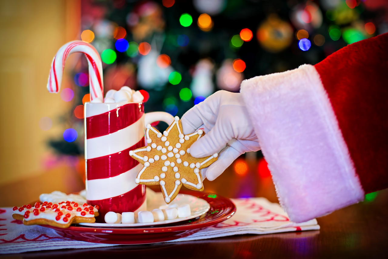 santa's arm picking up a snowflake cookie from a plate with a red and white stripped mug with a candy cane sticking out of it.