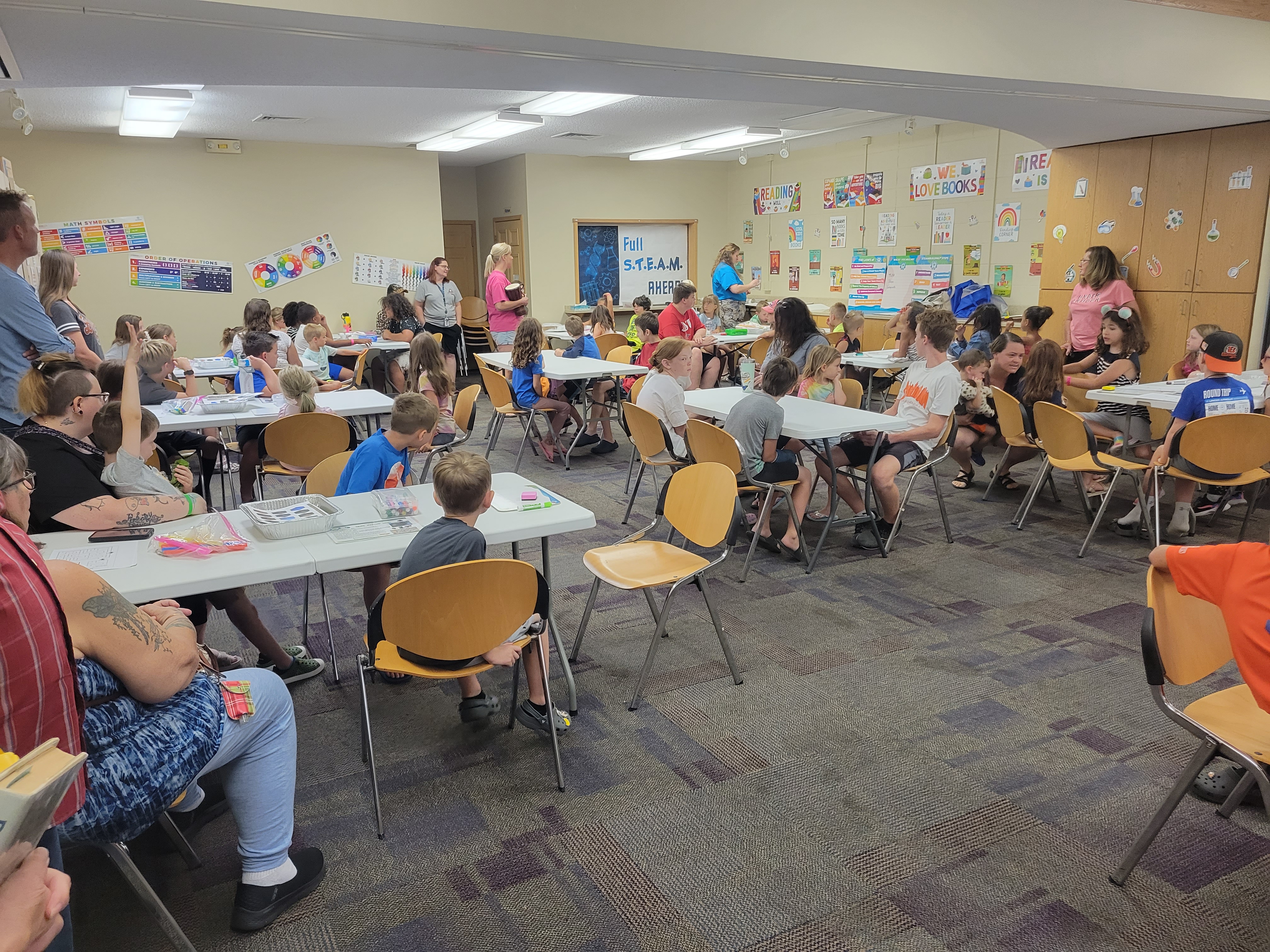 children and adults sitting around tables of supplies listening to instructions on how to build a marble maze.